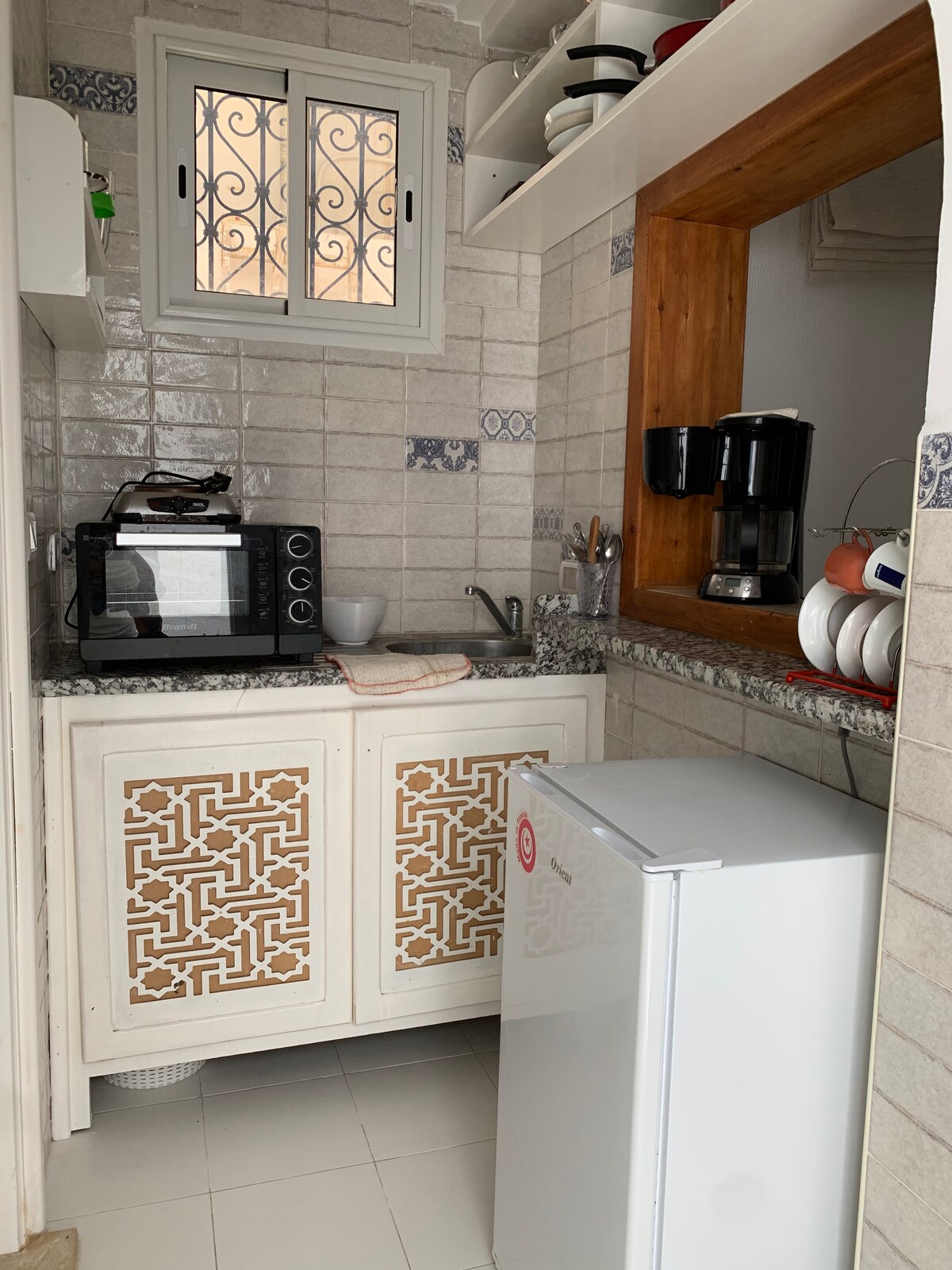 A compact kitchen features white and wooden cabinetry with intricate patterns. Modern appliances, including a microwave, coffee maker, and a spacious refrigerator, are present. A small sink is positioned beneath a window with decorative metalwork, allowing natural light to brighten the space.