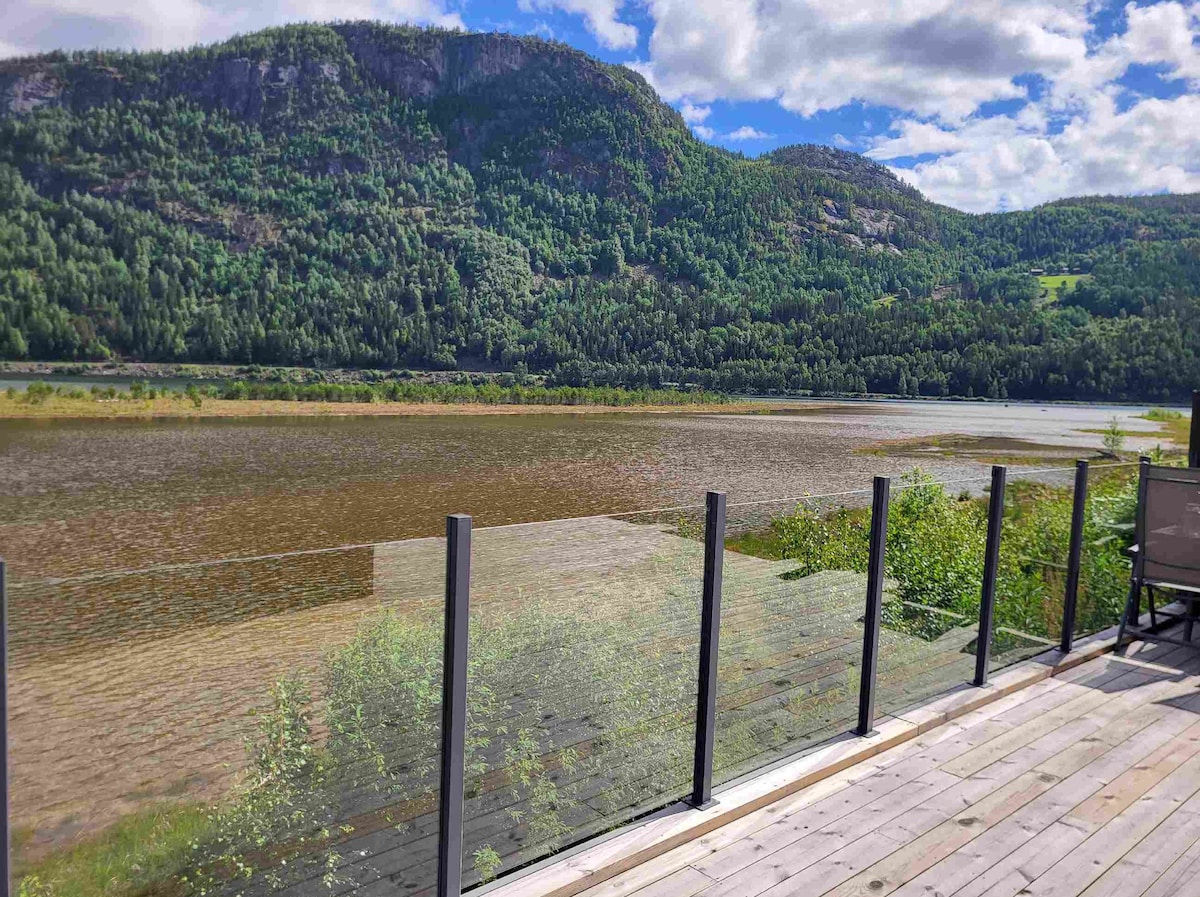 A spacious terrace features a glass railing overlooking a scenic view of lush green mountains and a calm fjord. The surrounding landscape displays a mix of greenery and water, contrasting against a bright, partly cloudy sky.