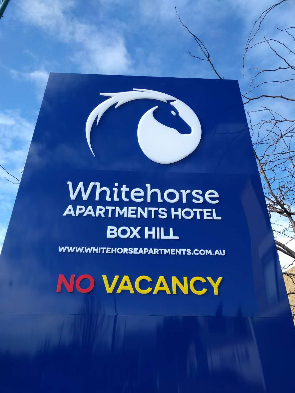 A prominent sign for Whitehorse Apartments Hotel features the hotel's name in stylish white lettering against a deep blue background. Below, the phrase 'NO VACANCY' is displayed in bold yellow letters, drawing attention. The sign is complemented by a graphic of a horse's head.