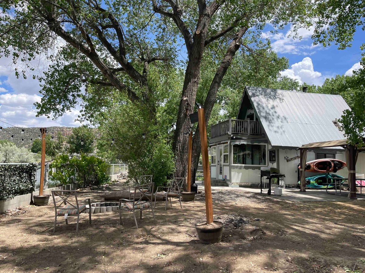 A spacious outdoor area features a large tree providing shade and multiple wooden chairs arranged around a fire pit. The cabin stands nearby, showcasing a peaked roof and a second-story balcony. Kayaks are positioned against the side of the cabin, ready for use.