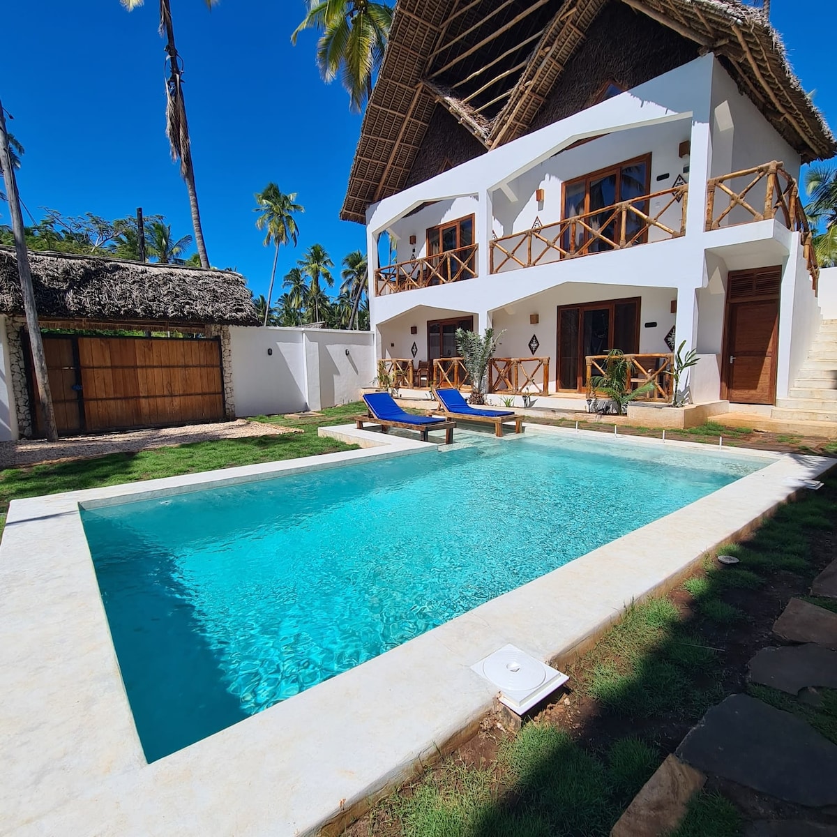 A relaxing open-air pool features crystal clear water, surrounded by a spacious lawn. The two-story resort building, with a traditional thatched roof, has private balconies and patio areas, offering views of the pool and nearby coconut trees. Sun loungers are positioned poolside.