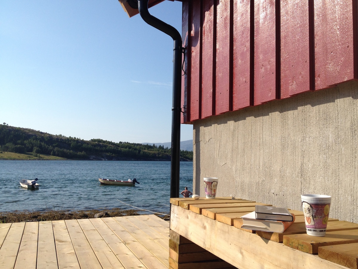 A wooden deck extends along the water's edge, featuring a low bench with several cups and a stack of books. In the background, two small boats are visible on the calm water, flanked by green hills under clear blue skies.