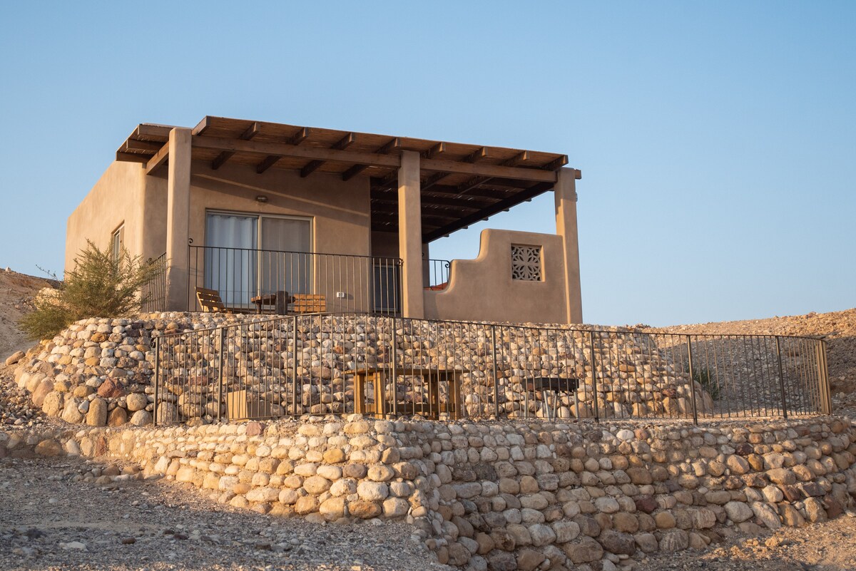 A cabin is perched on a stone terrace, featuring a covered porch with wooden seating. The building showcases a blend of natural earth tones and modern elements, with large windows allowing for ample natural light. The surrounding landscape highlights the desert's unique terrain.