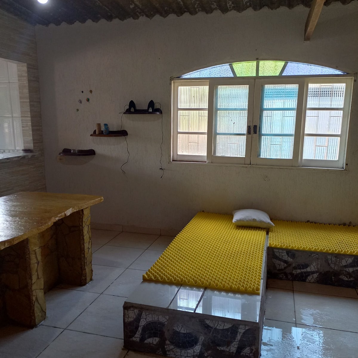 A simple interior space features a yellow foam sleeping area adjacent to a stone table. Natural light enters through a large window with colored panes, illuminating the room's textured walls and tiled floor. A small shelf and a pair of shoes are visible on the wall.
