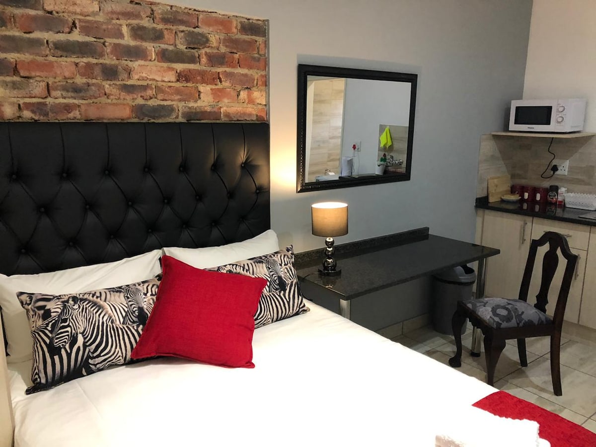 A comfortable sleeping area is highlighted by a tufted black headboard and stylish bedding. A small kitchenette is visible, featuring a microwave and a countertop. A framed mirror reflects the space, and a vintage chair adds an elegant touch.