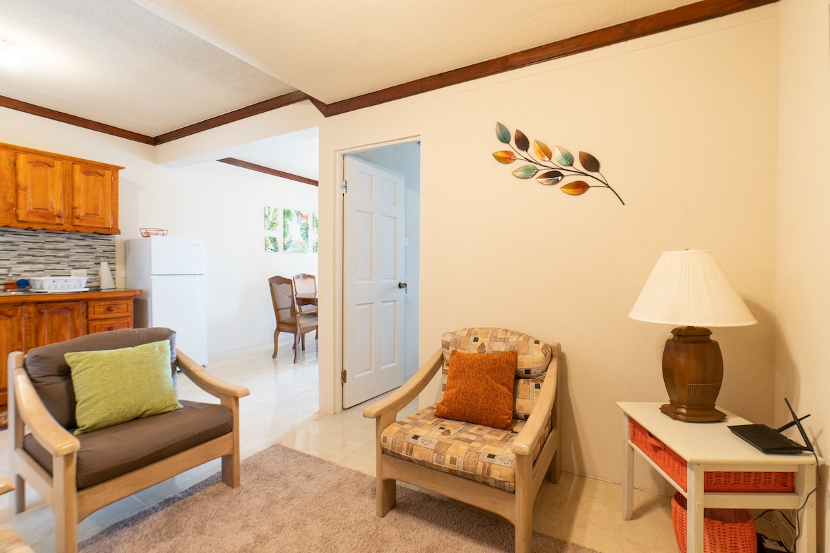 A living area is displayed featuring comfortable seating with accent cushions. A wooden table and chairs are visible in the adjacent dining area. Soft lighting is provided by a lamp on a small table, and decorative wall art adds a touch of color.