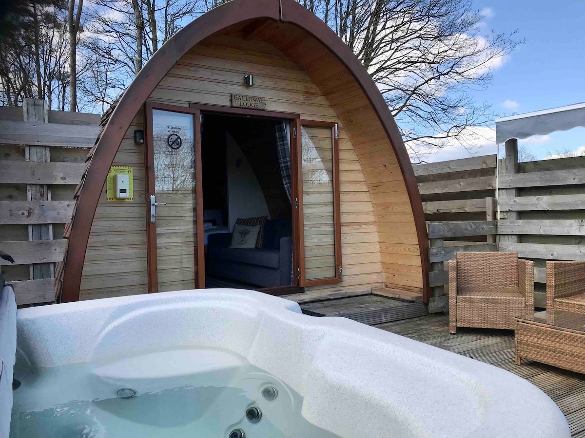An inviting lodge pod features a curved wooden exterior with a large entrance door. A cozy seating area is visible through the entrance. An electric hot tub sits in the foreground, surrounded by wooden decking and outdoor wicker furniture, under a clear blue sky.