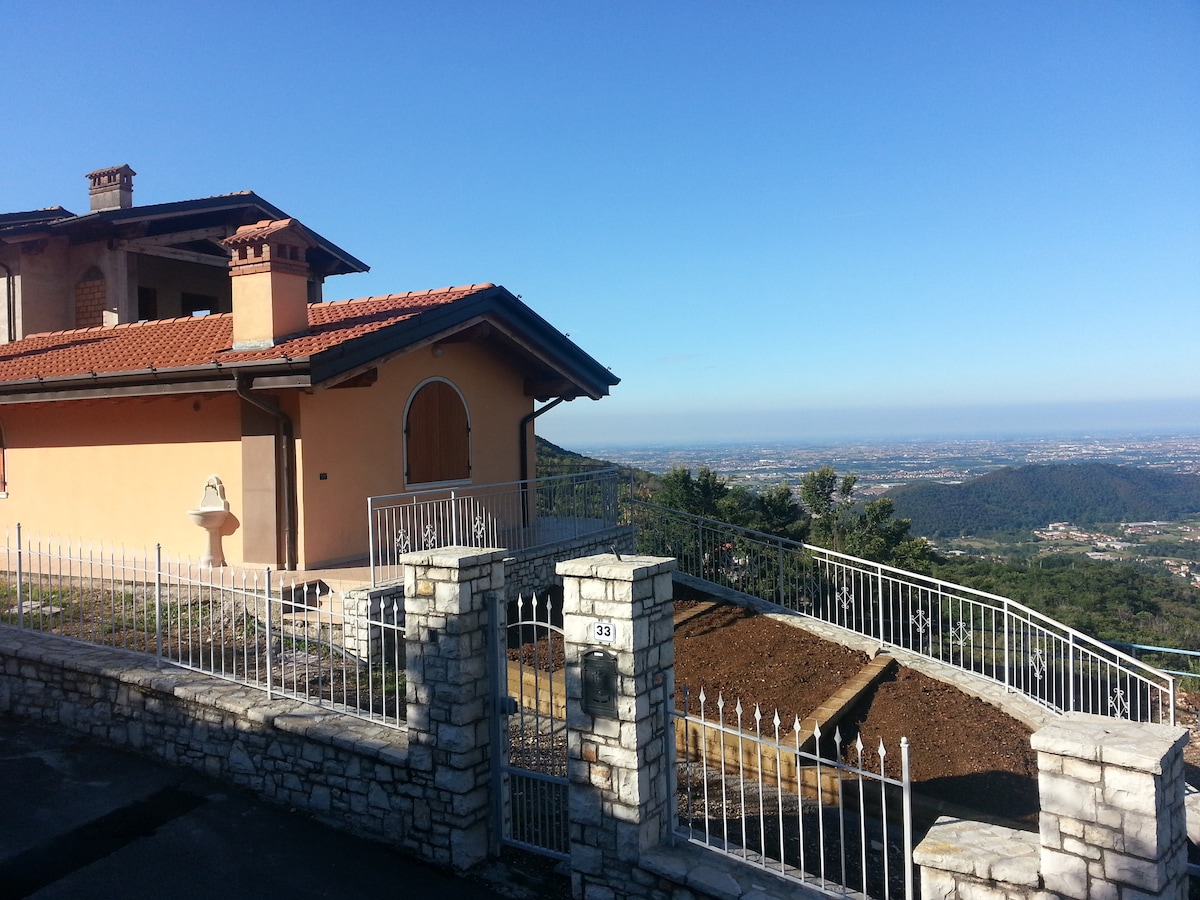 An independent house is shown with a light-colored exterior, featuring a terracotta roof. The surrounding landscape reveals a panoramic view of the valley below, set against a clear blue sky. A gated entrance with a stone wall frames the property.