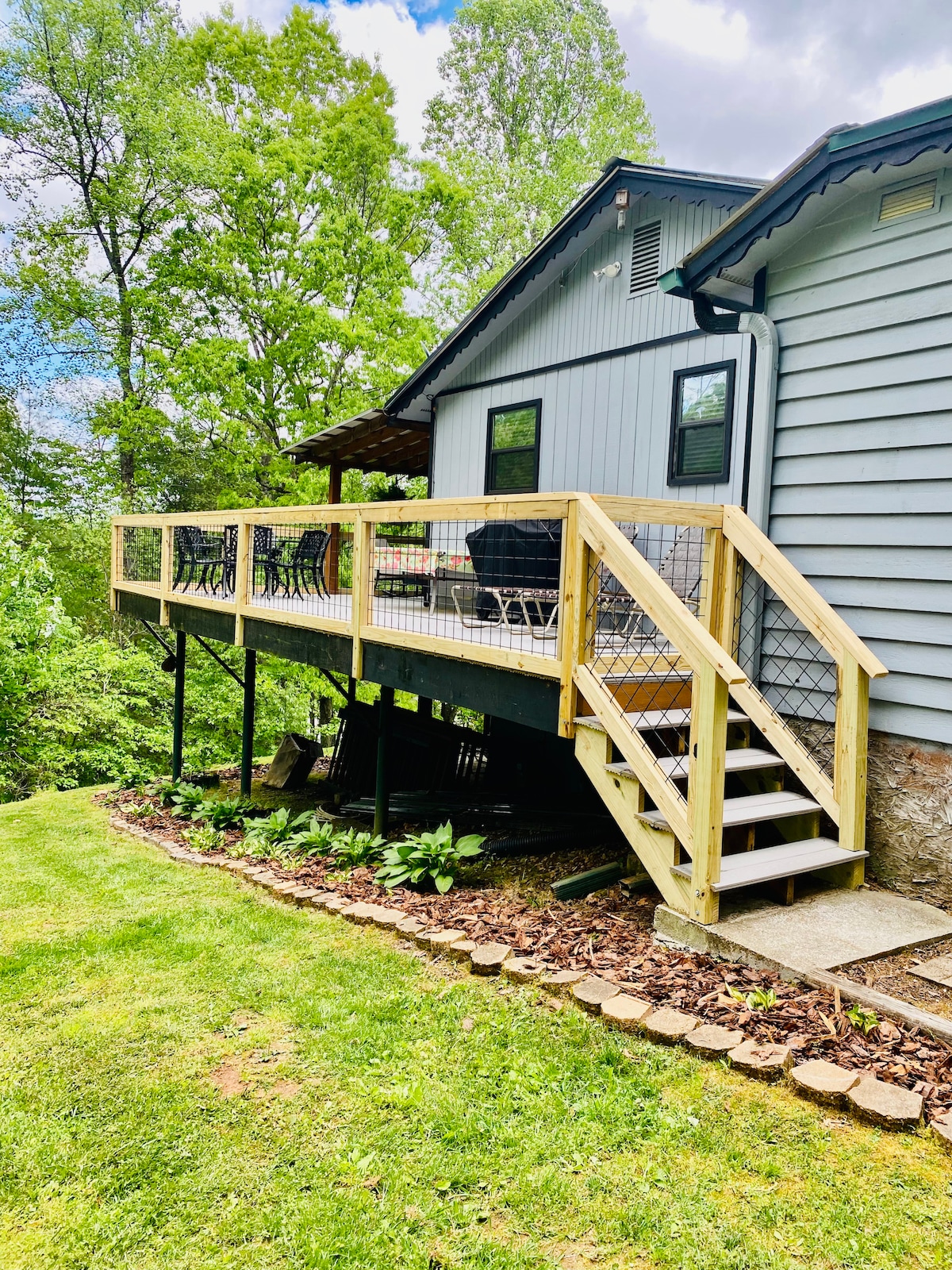 A spacious wooden deck is showcased, featuring a railing and steps leading down to a well-maintained yard. The deck is adorned with several black chairs, while lush greenery surrounds the area, enhancing the natural setting.