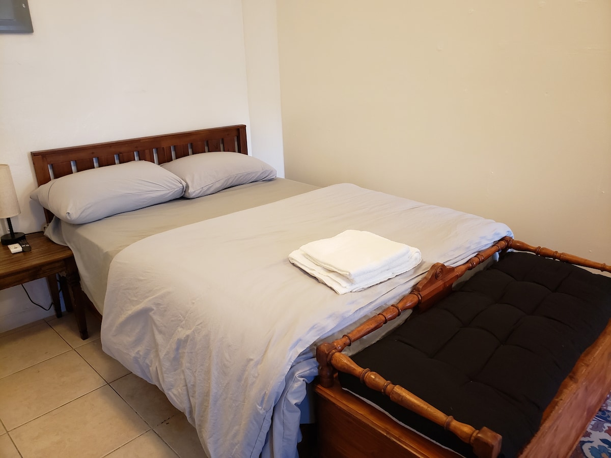 A spacious bedroom features a wooden bed with neatly arranged gray linens and two pillows. A small side table with a lamp is positioned beside the bed. A folded set of white towels is placed on the bed, and a smaller wooden bed is visible nearby.