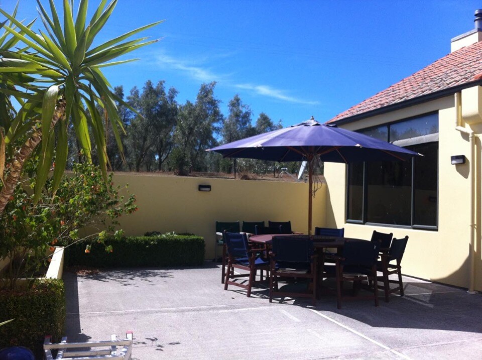 An outdoor seating area features a large shaded table under a blue umbrella, surrounded by green hedges. Several comfortable chairs provide ample seating for guests. The area is positioned next to the property, with a clear blue sky overhead and neat landscaping visible in the background.