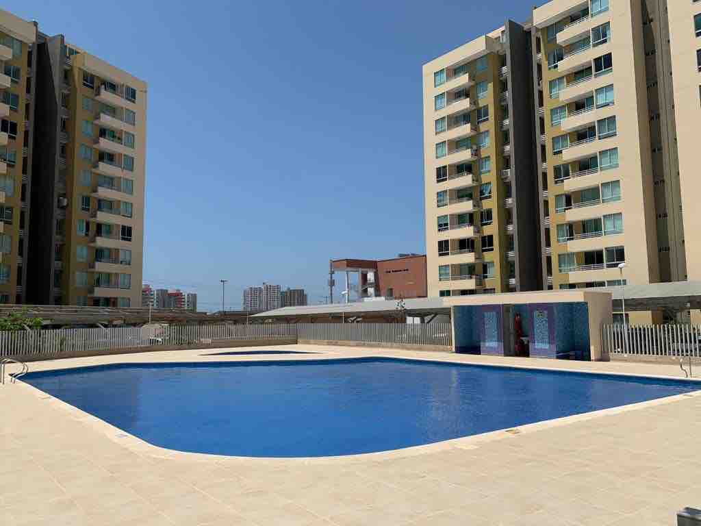 A large swimming pool occupies the center of the image, surrounded by a tiled deck. Multiple apartment buildings are visible in the background, featuring balconies and large windows, under a clear blue sky.