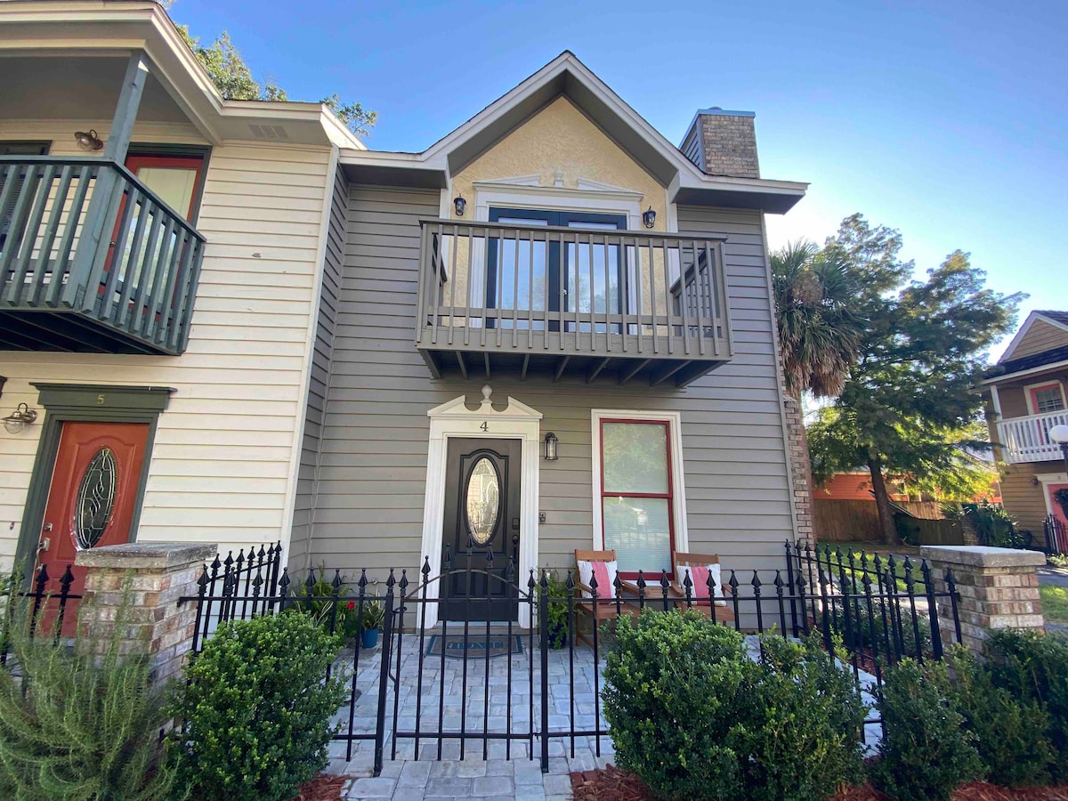 The exterior of a two-story townhome is shown, featuring a charming façade with a decorative balcony. A well-maintained courtyard is visible, surrounded by a low fence and landscaped with manicured shrubs, contributing to a welcoming presence.