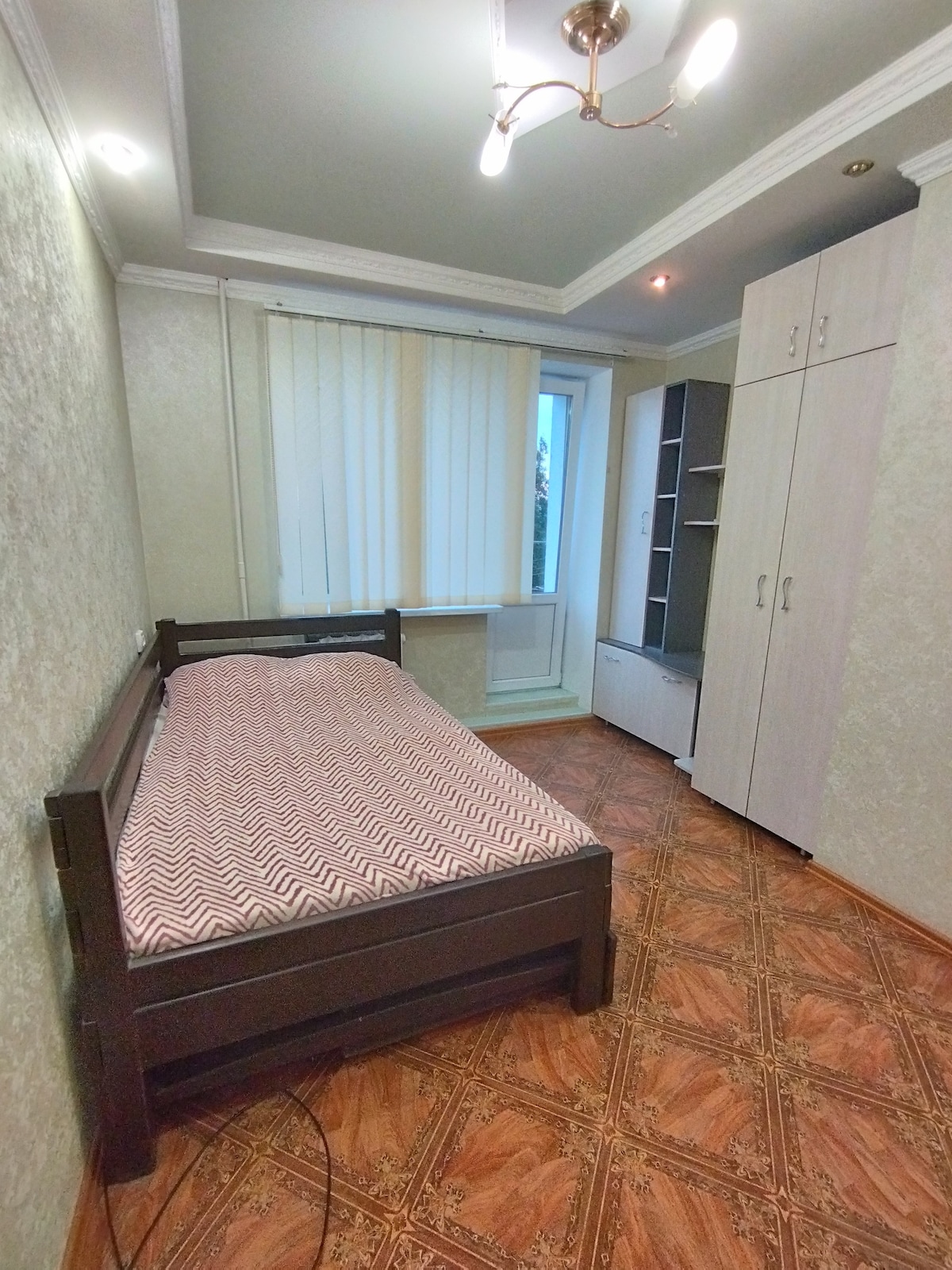 A compact bedroom features a single bed with a patterned blanket. Natural light enters through a window covered with vertical blinds. A tall wardrobe and shelving unit are positioned against the wall, providing ample storage space. The floor is adorned with a warm-toned patterned design.