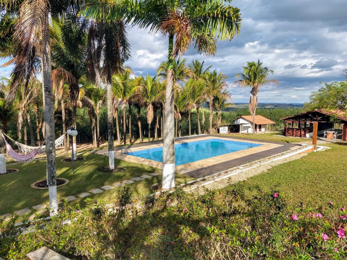 A spacious outdoor area features a crystal-clear swimming pool surrounded by palm trees. A patio with a roofed seating area and a hammock is visible nearby, with well-maintained gardens that include flower beds and natural grass, providing a tranquil setting.