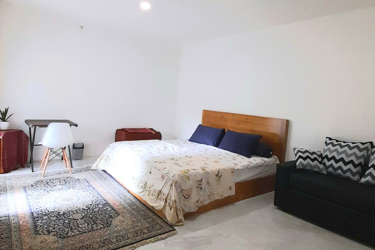 A bright room features a spacious bed dressed in a floral blanket with two dark pillows. A cozy area rug lies underneath, and a small table with an armless chair is positioned nearby. A dark sofa adds a comfortable touch to the space.