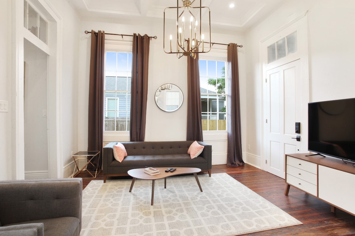 A well-appointed living area features a modern sofa complemented by accent pillows, a mid-century coffee table, and a flat-screen television. Large windows with flowing curtains allow natural light to fill the space, while the original hardwood floor adds warmth and character.