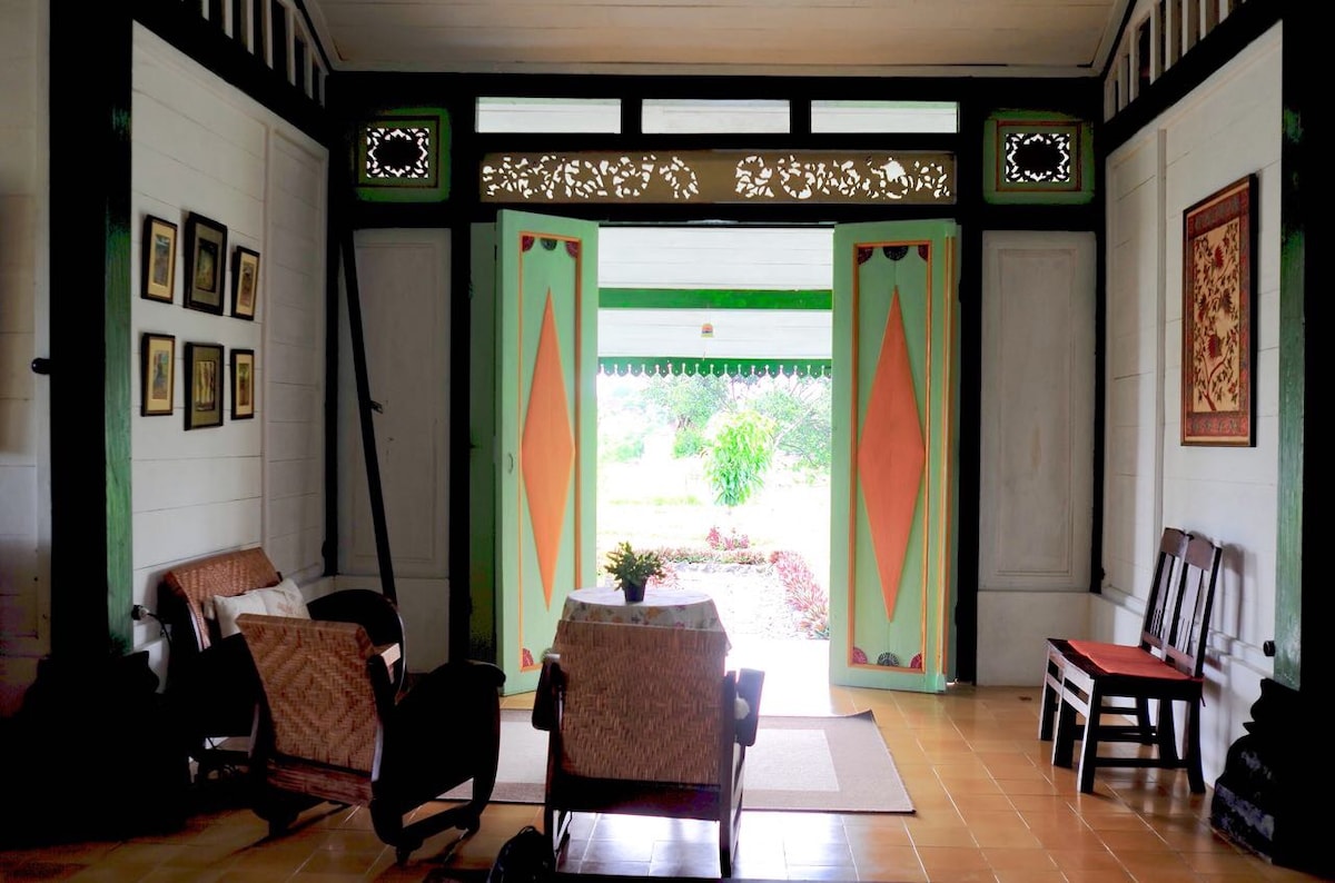 A welcoming entryway is framed by intricately carved doors painted in green and orange hues. Natural light filters in, illuminating a sitting area with woven chairs and a small table. The floor is tiled in a warm shade, contributing to the open, airy feel of the space.
