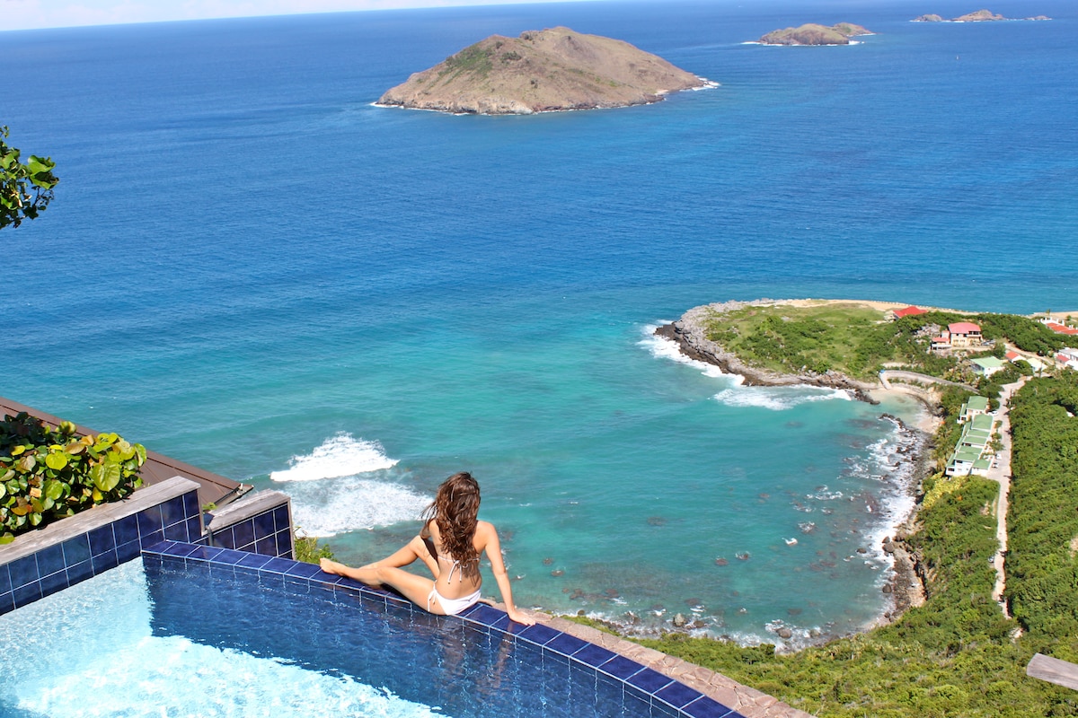 An infinity edge pool extends towards stunning ocean views, displaying a blend of turquoise and deep blue waters. Small islands and a coastal landscape are visible in the distance, creating a serene and picturesque backdrop.