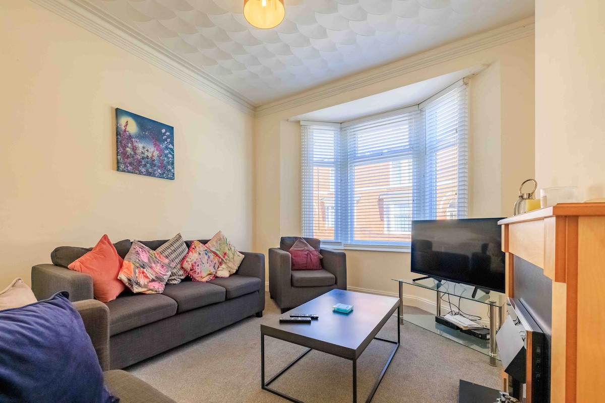The living room features a comfortable gray sofa and two matching armchairs, adorned with decorative cushions. A modern glass coffee table sits in the center, and a flat-screen TV is visible on a contemporary stand. Natural light filters in through large bay windows, illuminating the space.