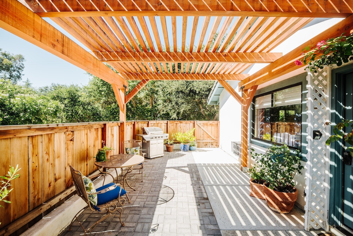 An inviting outdoor patio features a wooden pergola overhead, creating shade over a relaxing space. A grill is positioned to one side, complemented by a small table and two chairs, with vibrant greenery surrounding the area and enhancing the peaceful atmosphere.