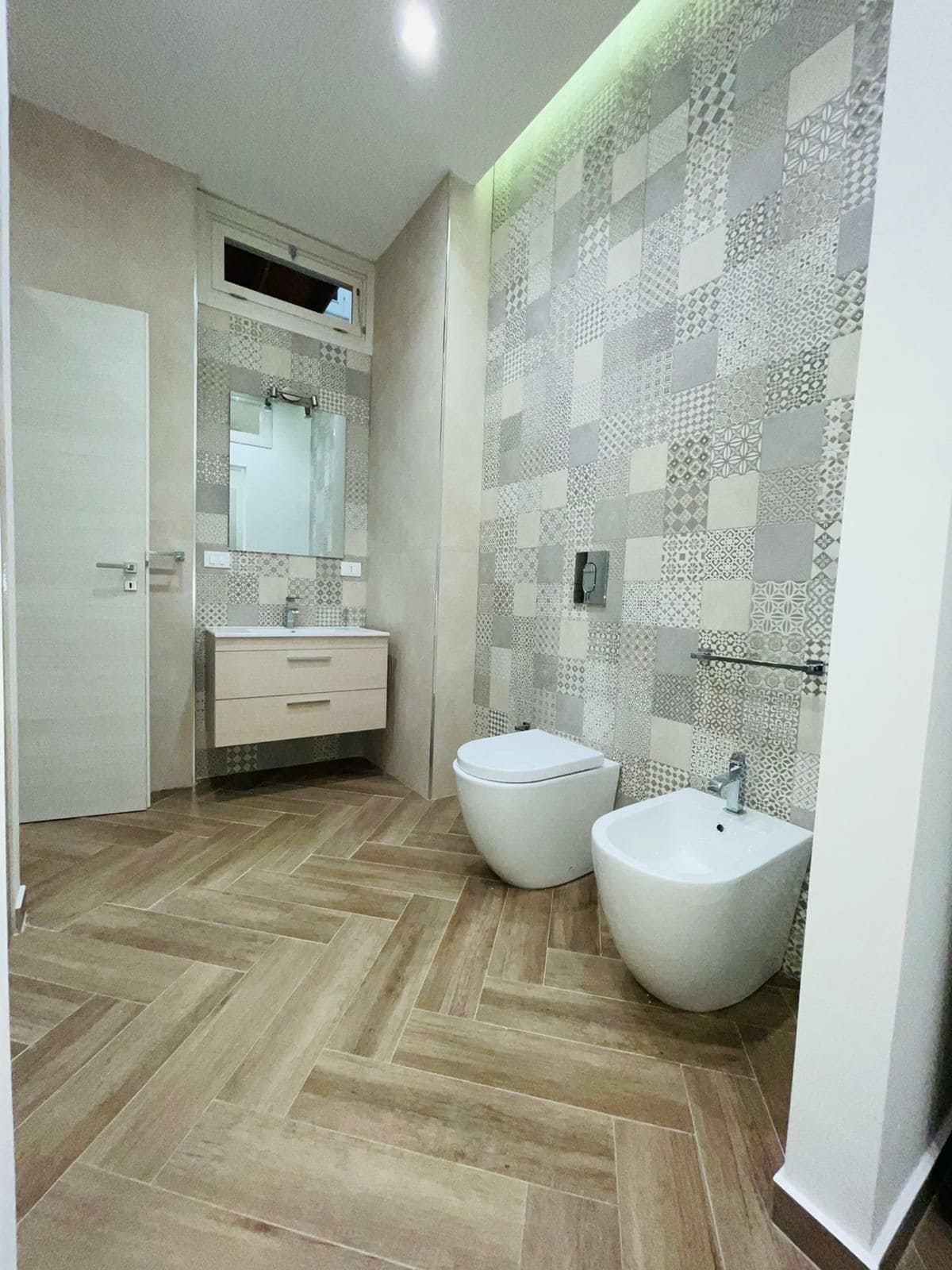 A modern bathroom is displayed, featuring a combination of patterned tile walls and wood-inspired tiles on the floor. A floating vanity with storage is present, alongside a toilet and bidet. The natural light is complemented by subtle green accent lighting.