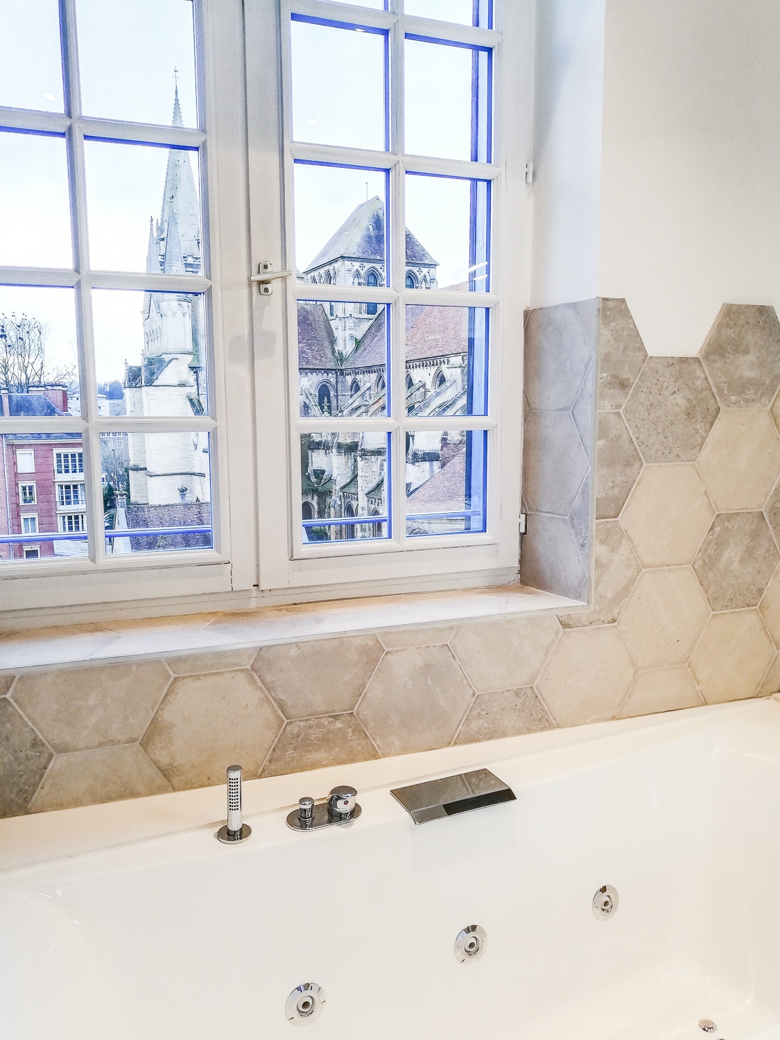 A modern bathroom features a large soaking tub beside a window, providing a view of the historical architecture outside. Hexagonal tiles decorate the walls, while a sleek faucet and shower controls are positioned on the tub's edge.