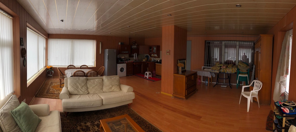 A spacious living area is showcased, featuring light wood paneling and large windows. A comfortable white sofa is visible, complemented by a decorative area rug. A kitchen area is present, containing appliances and a dining table with chairs, while additional seating options are offered.