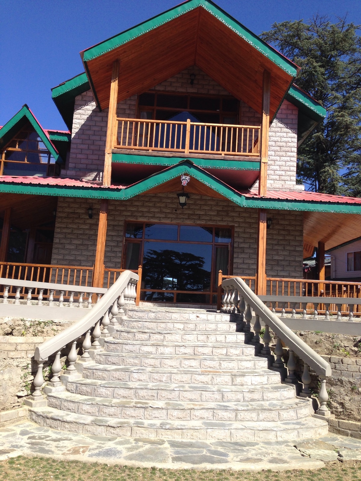 The entrance features a set of wide stone steps leading up to a wooden door, framed by large windows. The building showcases an inviting combination of stone and wood elements, topped with a green roof. Balconies extend from the upper levels, offering outdoor views.