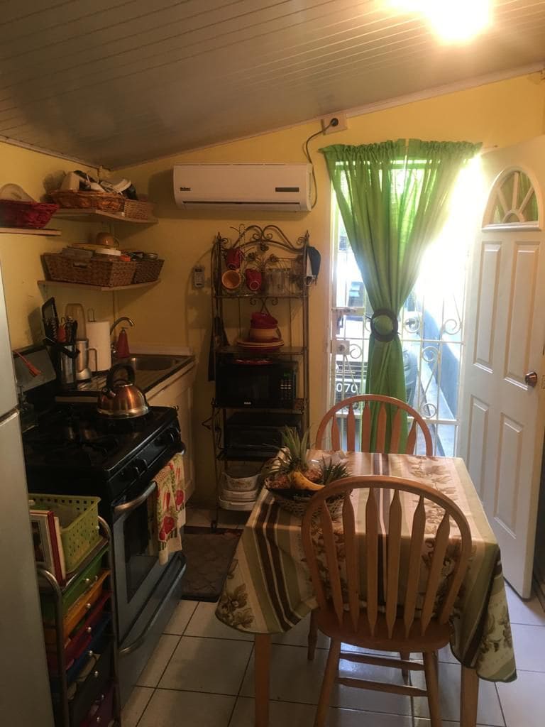A compact kitchen area is presented, featuring a round wooden table with chairs and a white tablecloth. Cooking appliances are visible, including a stove and refrigerator. Natural light enters through a window adorned with green curtains, complementing the yellow walls.