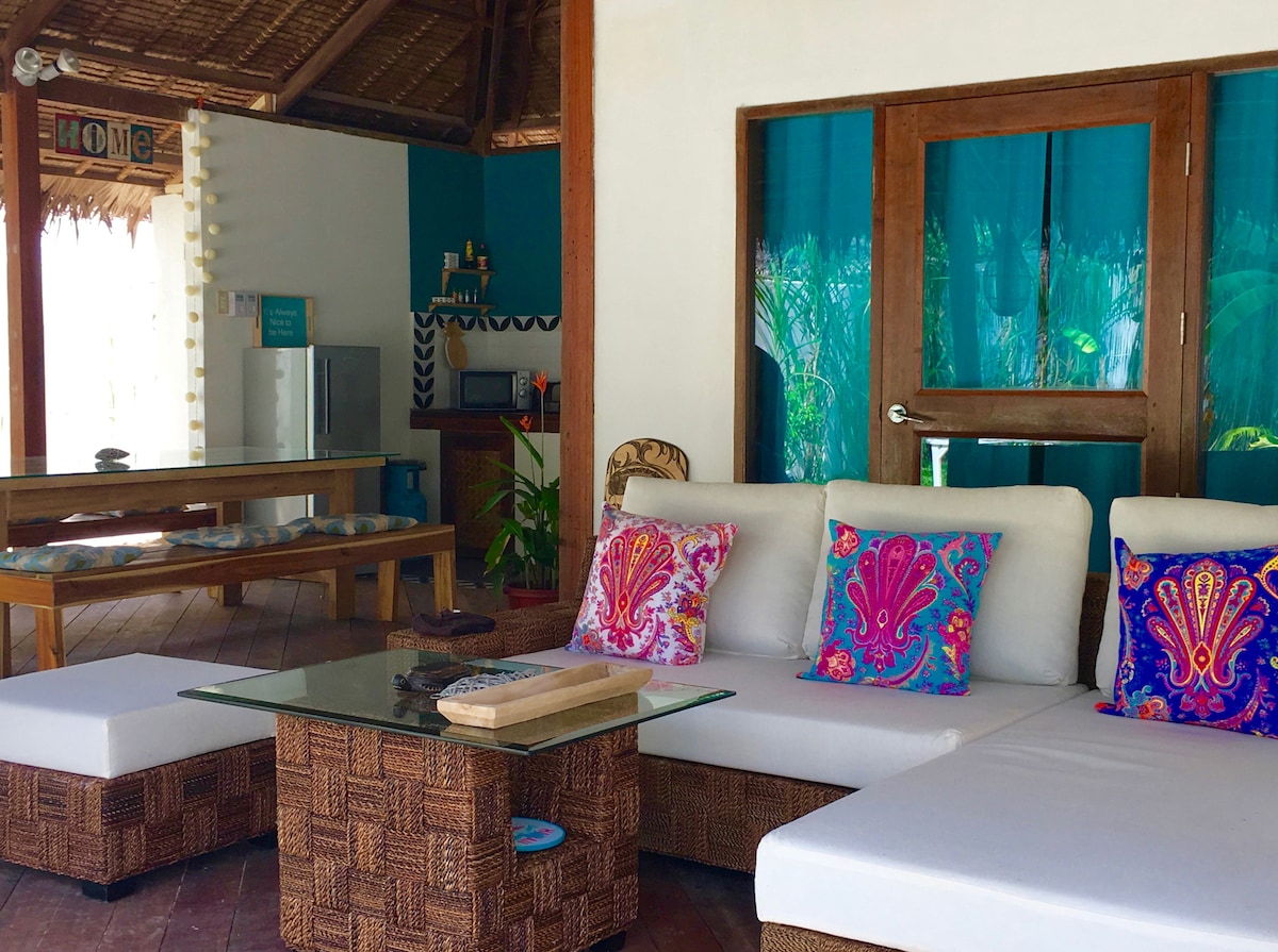An outdoor living area features a comfortable arrangement of light-colored seating with colorful decorative pillows. A glass coffee table sits in the center, surrounded by a rustic backdrop of wooden walls and vibrant blue accents. A fully equipped kitchen area is visible in the background.