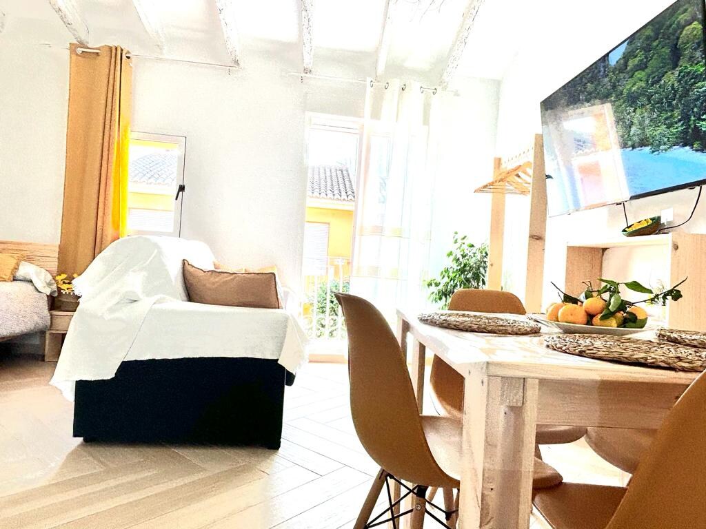 A bright, open-plan living area features a comfortable sofa draped with a light cover and a dining table surrounded by modern chairs. Large windows allow ample natural light, which highlights the wooden flooring and decorative elements. A mounted TV adds a touch of modernity.