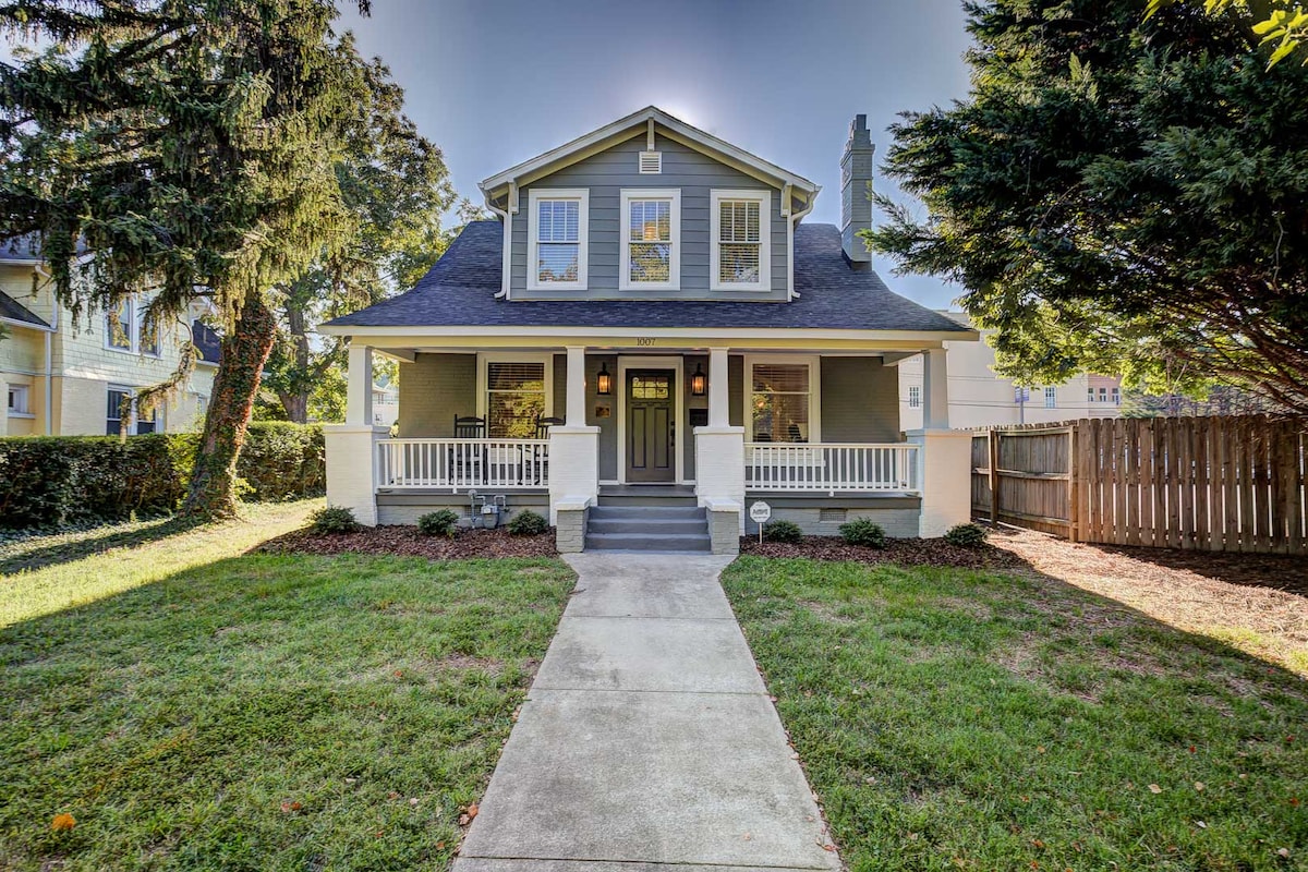 The charming exterior of a renovated Craftsman bungalow features a welcoming front porch with white railings and a gray door. A landscaped yard with green grass and lush shrubs is visible, bordered by a wooden privacy fence. Natural light highlights the structure under a clear sky.