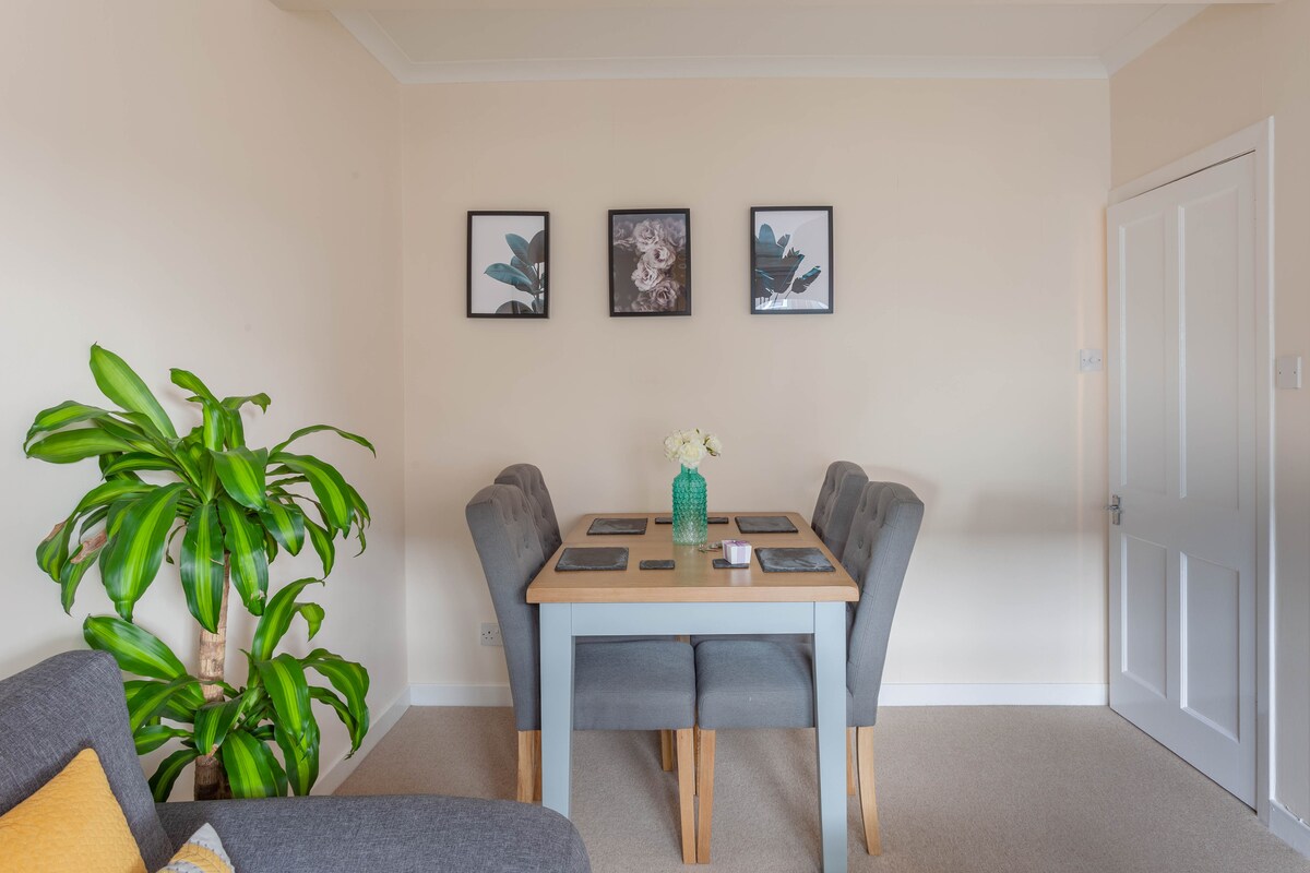 A dining area is presented with a wooden table surrounded by four grey upholstered chairs. Two framed prints are displayed on the pale wall above the table, while a green plant adds a touch of natural color to the space.