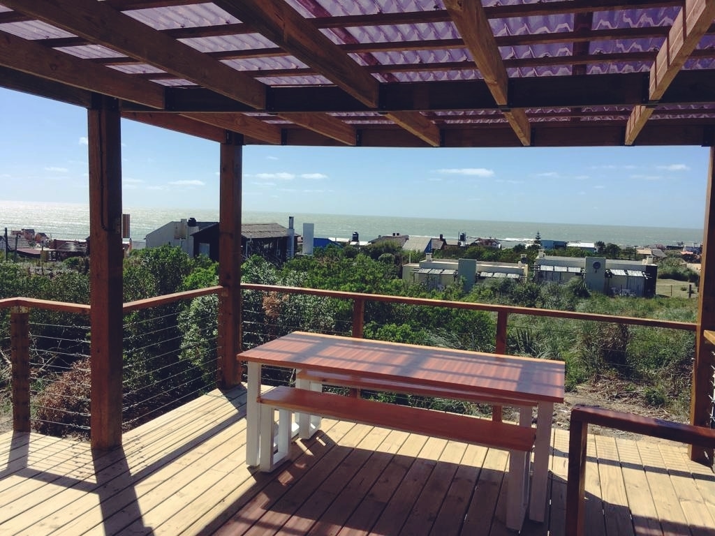 An expansive deck features a wooden table surrounded by greenery, offering unobstructed views of the ocean. The structure is partially covered with a polycarbonate roof that protects against UV rays, enhancing the outdoor space for relaxation and family gatherings.
