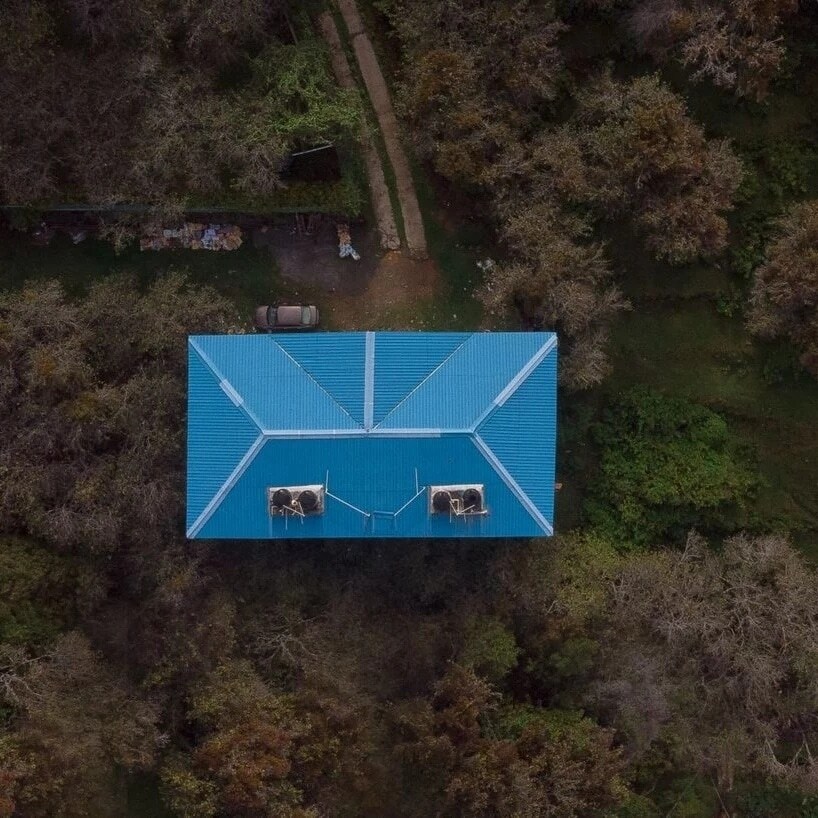 An aerial view of a cottage reveals a blue roof with two skylights. Surrounding the structure are trees with autumn foliage, blending greens and browns. A path is visible leading towards the cottage, providing a sense of connection to the natural landscape.