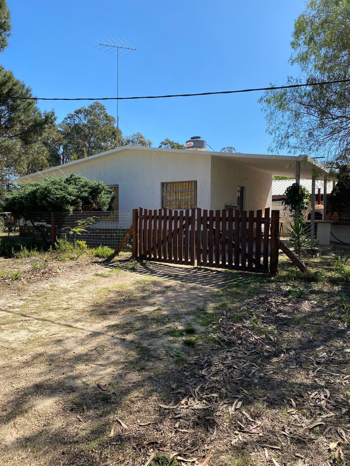 A single-story house is visible, framed by a lush green landscape. A wooden gate leads into the property, which features a spacious front yard and a mix of trees and shrubs. Natural light highlights the structure's clean lines and inviting entrance.