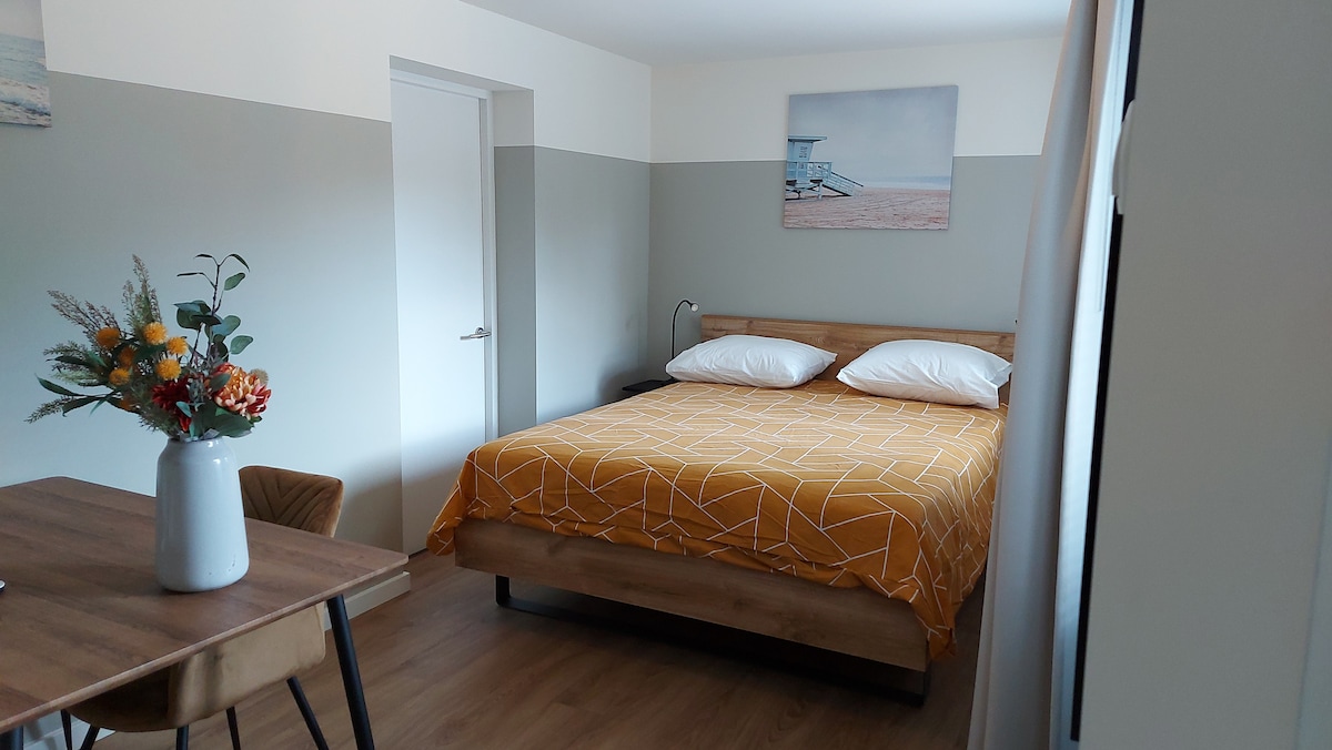 The bright bedroom features a spacious bed covered with a geometric-patterned yellow blanket. A wooden table with a chair is positioned nearby, complemented by a vase of flowers. Light-colored walls enhance the airy atmosphere, and a doorway leads to a separate hallway.