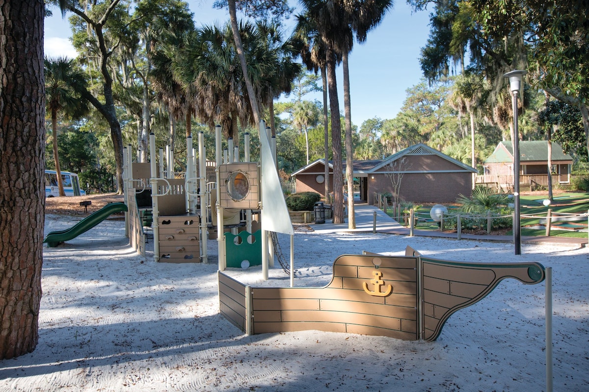 A children's playground is depicted, featuring a sandy area with a large ship-shaped play structure, complete with a steering wheel and climbing features. Surrounding palm trees and greenery provide a natural setting, while a slide and a nearby building are visible in the background.