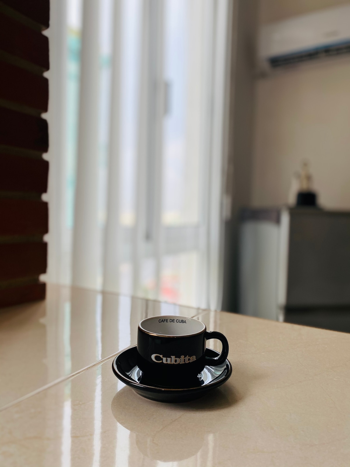 A black coffee cup labeled 'Cubita' is positioned on a matching saucer. Light filters through partially closed sheer curtains, illuminating the smooth countertop beneath. In the background, a glimpse of a refrigerator and an air conditioning unit is visible.