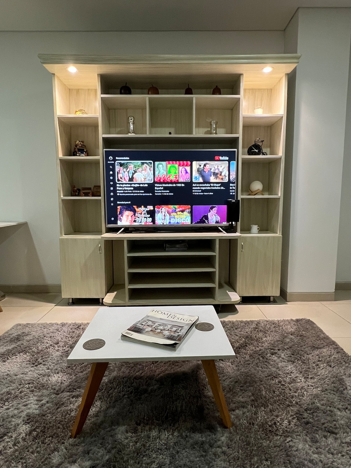 A modern entertainment center is showcased, featuring a large Smart TV mounted on an elegant light wood unit. Below the TV, a sleek media shelf is visible. The soft gray area rug adds comfort, while a coffee table with a magazine sits at the center.