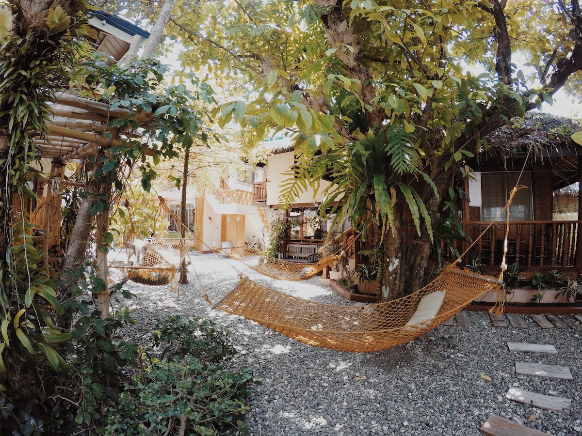 A serene outdoor area is framed by lush greenery and trees, featuring a hammock suspended between two supports. Gravel pathways meander through the space, with wooden structures and seating areas visible in the background, providing a tranquil retreat.