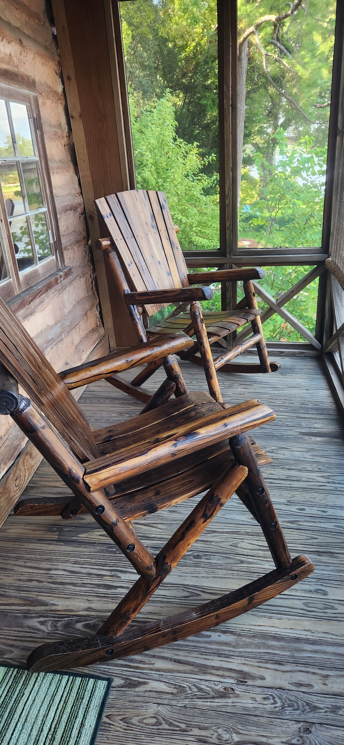 A screened-in porch features two wooden rocking chairs positioned on a wooden floor. Lush greenery is visible through the screens, providing a natural backdrop that enhances the serene setting.