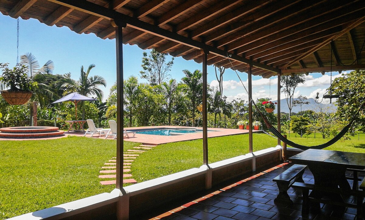 A spacious outdoor area features a hammock and a dining table, providing a comfortable gathering space. The view includes a well-maintained garden, a crystal-clear pool, and lounge chairs, framed by lush greenery and distant mountains under a clear blue sky.