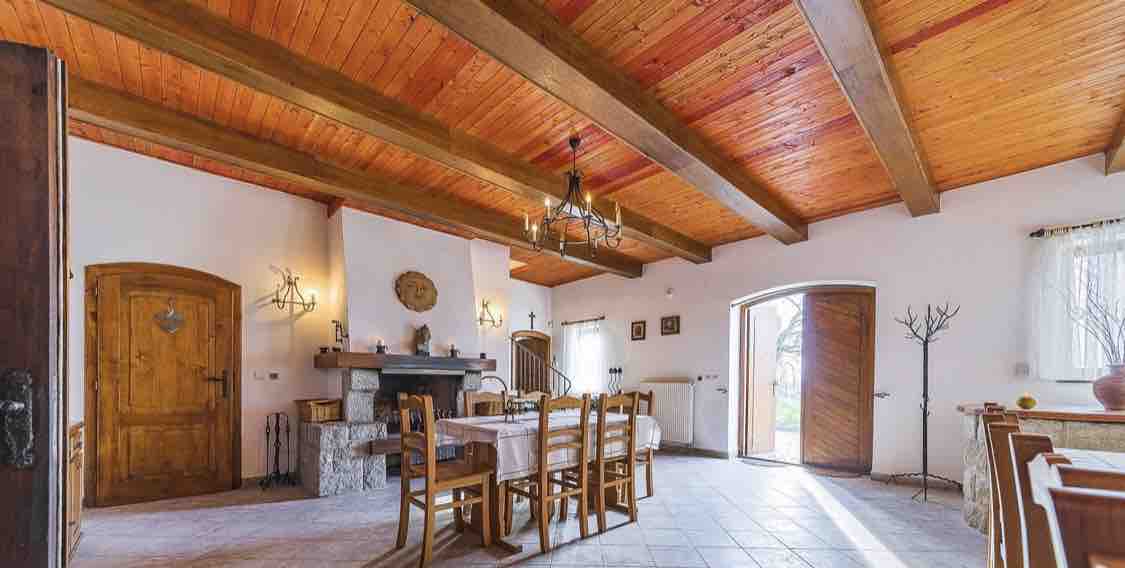A spacious communal area features a wooden ceiling with exposed beams, illuminated by natural light from large windows. A central fireplace is positioned against the wall, surrounded by wooden furniture. A dining table is set with several chairs and decorative wall elements add character.