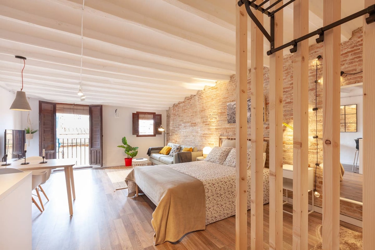 An open-plan loft features exposed brick walls, creating a textured backdrop for the space. A comfortable double bed is positioned near a bright window, while a sofa invites relaxation. A small dining area with a table and chairs is nearby, offering functionality within the layout.