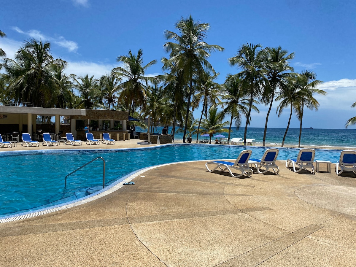 A large pool area is featured, surrounded by numerous lounge chairs arranged under swaying palm trees. The pool reflects the clear blue sky, with a distant view of the ocean enhancing the setting. A small restaurant is visible nearby, providing convenient access for guests.