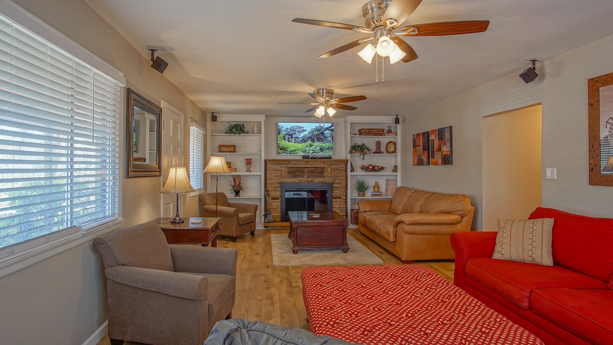 A comfortable living area is furnished with a brown leather sofa, a red couch, and a gray armchair. A coffee table sits in front of a rustic stone fireplace, accompanied by shelving displaying decorative items. Ceiling fans above provide airflow, and natural light enters through the window.