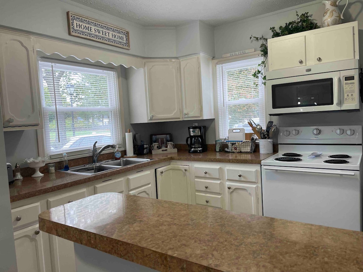 The kitchen features light-colored cabinetry and a countertop accented by a backsplash. Natural light enters through two windows, illuminating the workspace. Appliances include a microwave and a stove, while a coffee maker and various utensils are neatly arranged on the countertops.