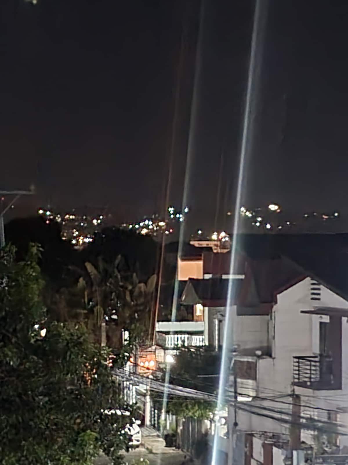 The night view captures a city skyline adorned with shimmering lights, set against a dark backdrop. Nearby residential structures and foliage frame the scene, providing a contrast to the illuminated buildings in the distance.